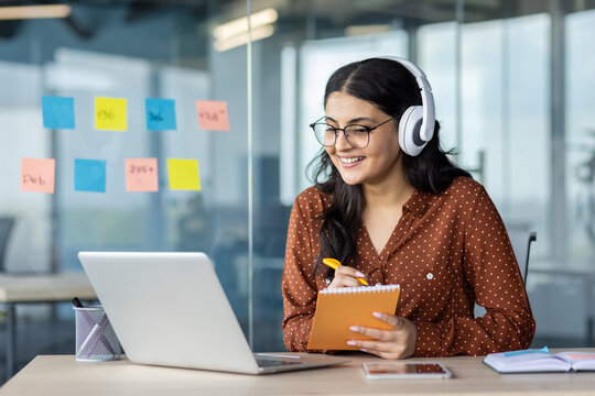 Woman watching online video course, training conference inside office at workplace. Office worker in headphones using laptop for remote meeting, writing data in notebook.