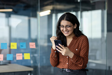 Successful woman at workplace standing with phone in hands. Businesswoman celebrating victory and...