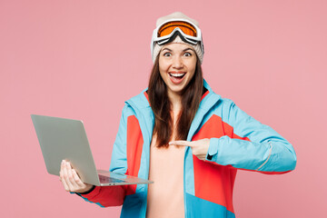 Skier young IT woman in blue padded jacket ski goggles mask spend extreme weekend winter season in mountains hold use work point on laptop pc computer isolated on plain pink background Hobby concept