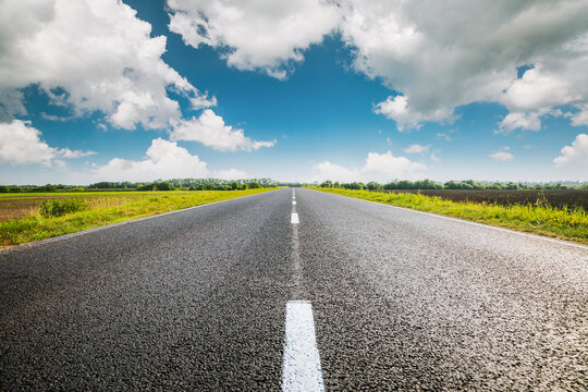 A straight asphalt road passing through agricultural fields under a blue sky with white fluffy clouds stretching into the distance.