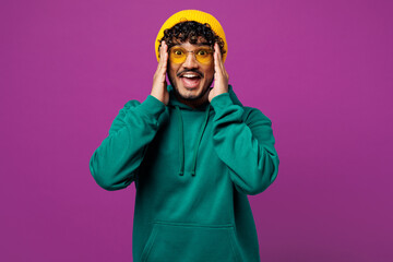 Young happy shocked surprised excited Indian man he wear green hoody yellow hat sunglasses casual clothes hold head look camera isolated on plain purple background studio portrait. Lifestyle concept.