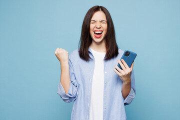 Young happy Caucasian woman she wears shirt casual clothes hold in hand use mobile cell phone do winner gesture isolated on plain pastel light blue cyan background studio portrait. Lifestyle concept.