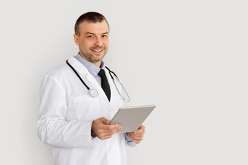 Medical Care And Technology Concept. Portrait Of Smiling Confident Doctor Using Digital Tablet, Standing Isolated On White Studio Background, Copy Space. Physician wearing coat and stethoscope