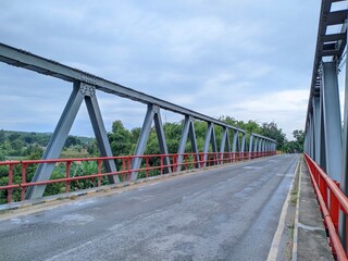 Naklejka premium A long, steel truss bridge stretches across a river or valley. Lush greenery lines the horizon. The bridge has red railings and a paved road runs along its center.