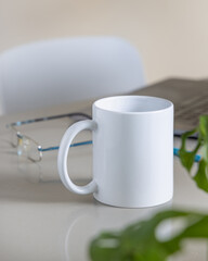 White coffee mug on grey office table against laptop, glasses and plant closeup, mockup