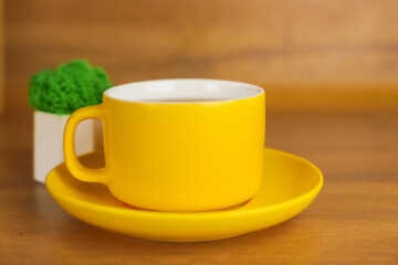 A yellow mug with a saucer, in which a hot drink is poured, stands on a wooden background, behind it you can see a plant in a pot view from the side. The concept of tea drinking, tea traditions. High