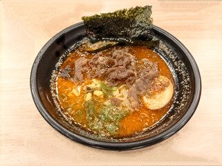 Japanese cuisine. Ramen noodle soup with sliced beef, egg, chives, sprouts and a piece of seaweed in a black bowl on an isolated wooden table.