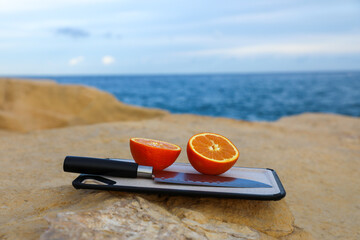 On a rocky seashore on a cutting kitchen board, next to a kitchen knife against the backdrop of the sea, lies a juicy, ripe, appetizing orange, cut in half.