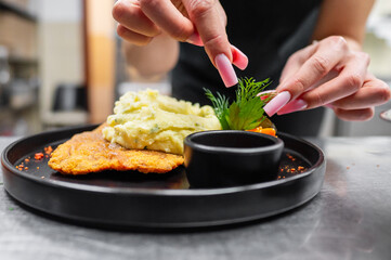 A close-up of a person serving creamy mashed potatoes over a crispy fried dish, placed beautifully on a black plate. The scene captures a moment of culinary preparation and enjoyment.