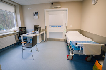 A clean hospital room with a bed, desk, chair, computer, and door labeled “Uzgaidāmā Telpa,” featuring bright lighting and a window view..