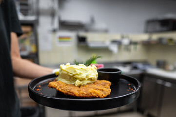 A person serves a black plate featuring crispy fried chicken alongside creamy mashed potatoes topped with herbs, in a cozy kitchen setting. Ideal for food-related content.