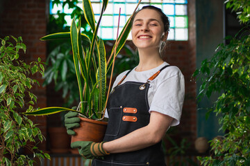 Running of own business. Young woman florist holding plant in pot wearing apron in botanical store. Happy small business owner working at flower shop smiling surrounded by plants. Small business