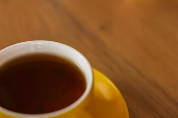 A yellow mug of black tea sits on a saucer in the lower left corner of the photo on a wooden background with space for the dough. The concept of tea drinking, break. High quality photo