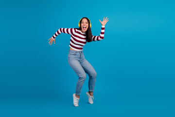 Young woman dancing happily in striped shirt and blue jeans against vibrant blue background