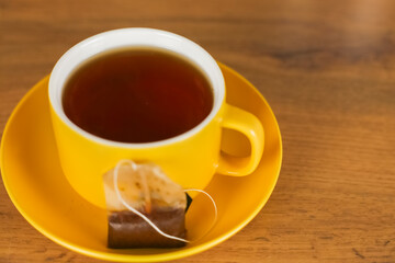 Side view of a yellow cup in which hot black tea is poured, which stands on a yellow saucer and on it lies a tea bag on a wooden background on the left with space for text. Concept of tea drinking