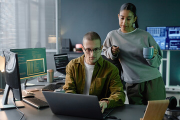 Medium shot of senior male programmer with glasses using laptop reviewing code while helping female junior developer collaborating at working desk in office