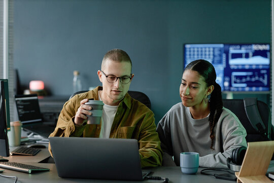 Medium shot of female IT programmer discussing with female colleague code debugging while collaborating over cup of coffee at working desk with laptop in office