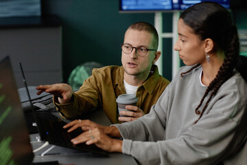Portrait shot of male IT programmer with glasses pointing at laptop screen while consulting female colleague during discussion at work meeting in office