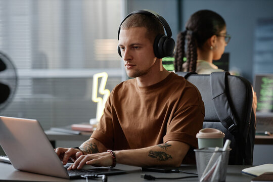Medium shot of confident male IT developer working in headphones immersed in coding on laptop at cluttered desk in corporate office, copy space