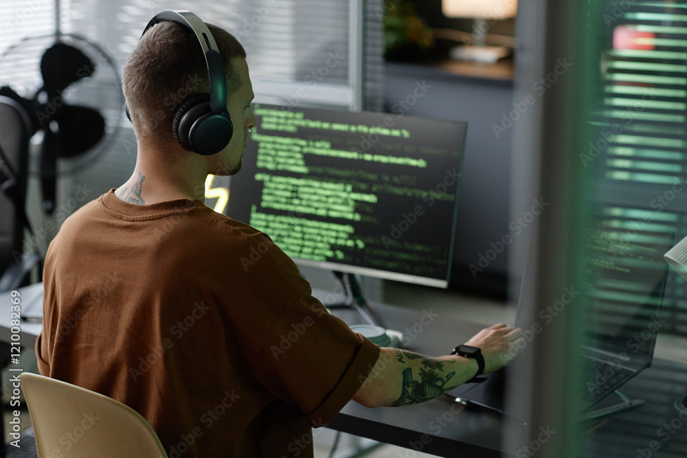 Poster side view shot of male programmer in headphones writing code on laptop looking at multiple screens w