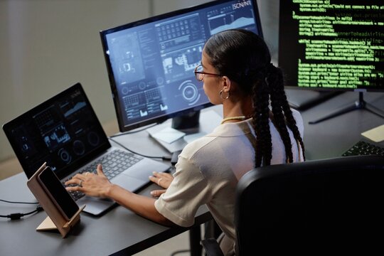 Side view of female programmer with braided hairstyle sitting at professional workstation equipped with multiple screens using laptop during long working day in office