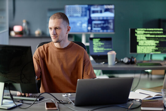Medium shot of calm male programmer typing on computer keyboard working alone at cluttered desk with laptop, captured against screens displaying lines of code in tech company office