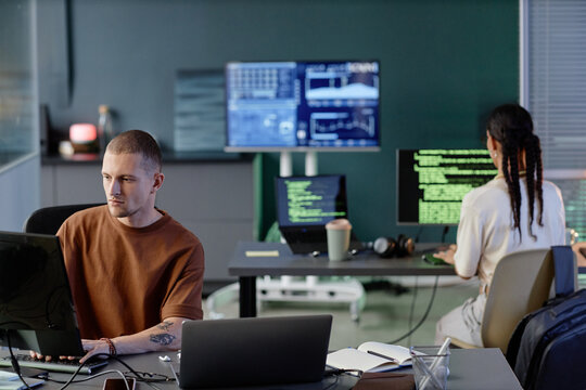 Wide angle shot of male programmer coding on computer at desk working with colleagues in spacious room of modern IT company office, captured against lots of screens with code lines