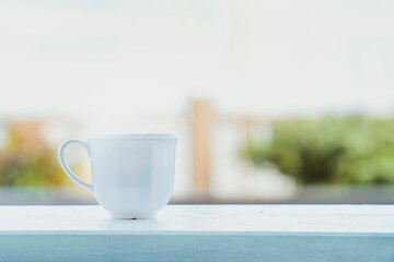 Cozy white coffee cup with steam on a wooden railing, bright morning scene with natural blurred background
