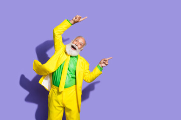 Portrait of funny extravagant aged man point fingers empty space ad yellow suit isolated on purple color background