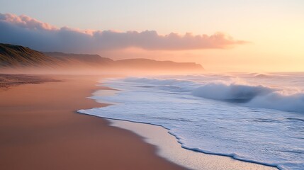 Obraz premium Sunrise Coastal Serenity: Waves gently caress a pristine sandy beach at sunrise, the soft golden light illuminating the misty coastline creating a breathtakingly serene atmosphere.