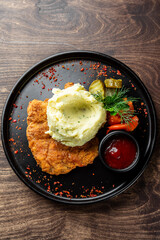 A delicious serving of crispy fried chicken with creamy mashed potatoes, accompanied by a side of mixed vegetables, pickles, and a small bowl of ketchup, all on a black plate.