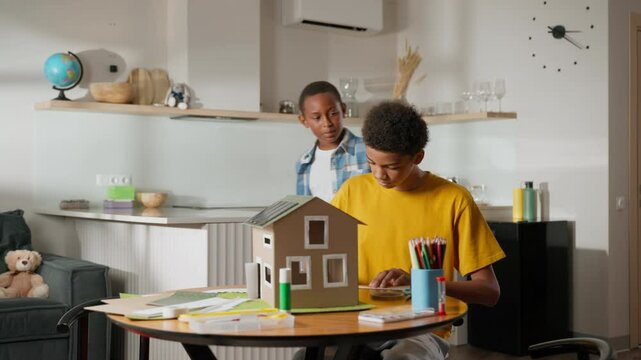 Older brother is already sitting at table, starting work on solar-powered model house. Younger brother enters frame, approaching table to join school science project. Sets collaboration for teamwork - Powered by Adobe