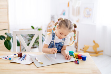 a little child girl paints a drawing with a brush in an album and smiles, a happy child draws a...