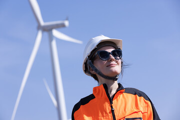 Engineer woman in hardhat with laptop and blueprint working on wind turbines at electricity power station background. Renewable energy, wind turbine generate electricity to produce energy .