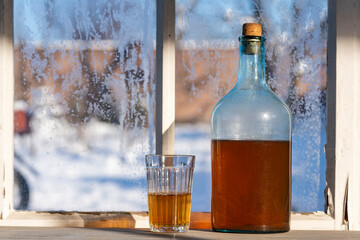 Big bottle with a drink made from fermented birch sap on the windowsill on winter sunny day. Traditional Ukrainian cold barley drink kvass in a glass jar and glass on table near yard
