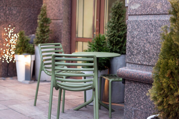 Fototapeta premium An inviting outdoor cafe area featuring green metal furniture, potted evergreens, and warm decorative lights. A serene and modern setting for relaxation and enjoying a peaceful urban atmosphere.