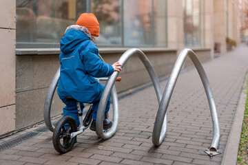 A young child dressed warmly explores an outdoor space while riding a small bike. The child interacts with an urban bike rack. Curiosity, activity, and discovery in a modern city environment.