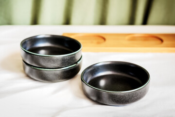 Black ceramic bowls arranged on a white tablecloth with a wooden tray and green fabric in the background