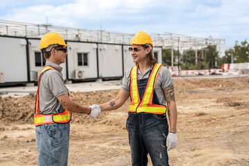 Two construction workers wearing hard hats and safety vests shaking hands at a construction site, symbolizing collaboration and successful teamwork with modular units in the background.