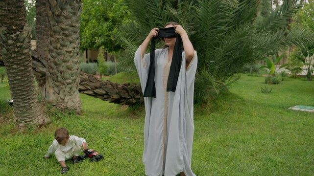 Mother playfully ties blindfold over her eyes and reaches out to catch her son in lush garden of family estate. Boy laughs and dodges her arms, creating joyful moment filled with fun and bonding