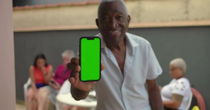 One Elderly black man holding a smartphone with a green screen, smiling confidently, showcasing technology outdoors, with diverse adults interacting in the background