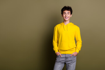 Young male wearing a yellow hoodie smiling confidently against a khaki background with hands in...