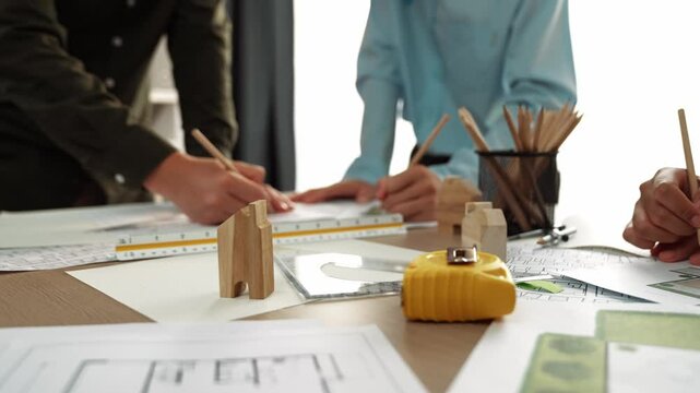 Smart architect and safety engineer discussion about building blueprint on table with architectural document, measurement tools and wooden block scatter around at modern office. Closeup. Delineation.