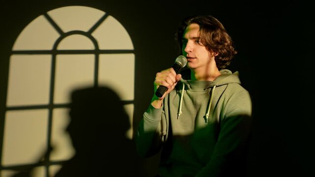 Portrait of man in green neon singing in karaoke microphone, standing in spotlight isolated on dark wall with window pattern studio background.