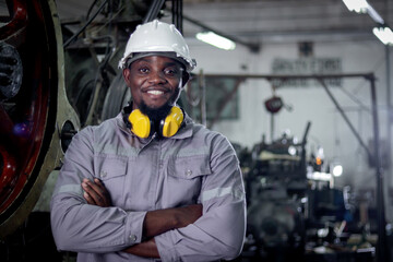 Portrait of African industrial engineer man with protective workwear and safely helmet stands with arms crossed at manufacturing factory. Smiling male worker working at steelmaking metal industry. © Stella