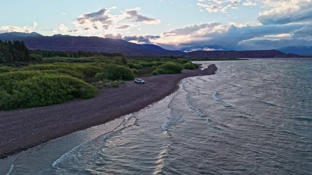 The end point of the journey.This captivating drone footage reveals a pristine lake, a lone white 4x4 parked on its shores, and the dramatic backdrop of green trees and the majestic Andes Mountains. 