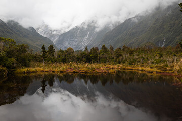 Misty reflections on small lake with snow-covered Franz Josef Glacier in the background and big clouds rolling over mountains (South Island, New Zealand)