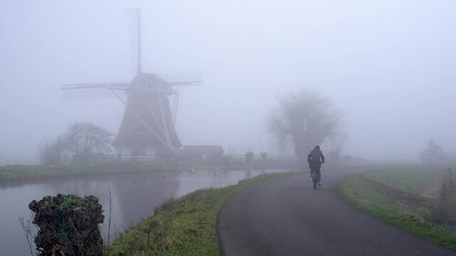 A Biker in Dutch Countryside Passes a Traditional Windmill, Cycling in
The Netherlands in Misty Weather With Low Visibility