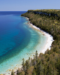 A lookout view of the deep blue and turquoise waters of a rocky shoreline, cliffs, and a cape point, along lush green forests. 