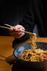 Close-up of a person using chopsticks to lift wok noodles from a steaming bowl of wok